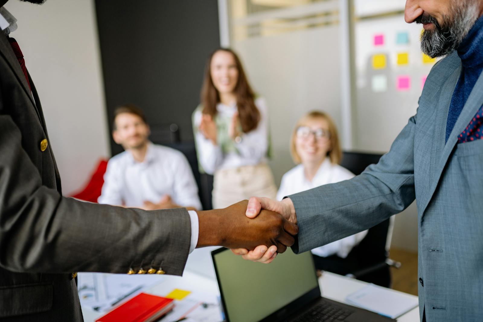 Zwei Geschäftsleute geben sich nach einer Einigung im Büro die Hand, Kolleginnen und Kollegen applaudieren, als Symbol für gewonnenes Vertrauen und eine Investitionszusage